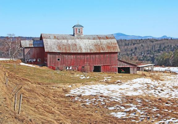A weathered old red barn stands in a field of dried grass and patches of snow with blue mountains in the distance.