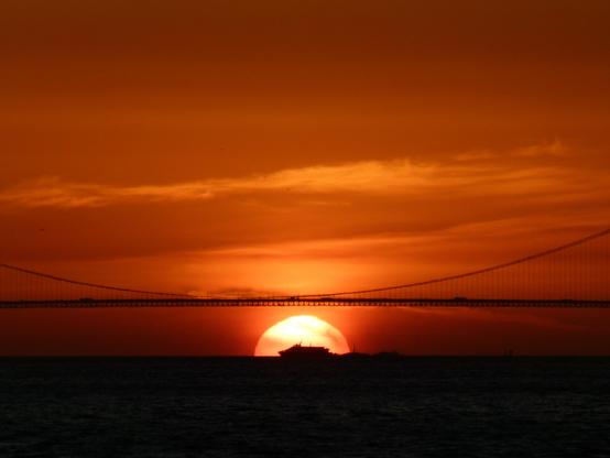 a ferry silhouetted by the setting sun in the center of the Golden Gate Bridge
