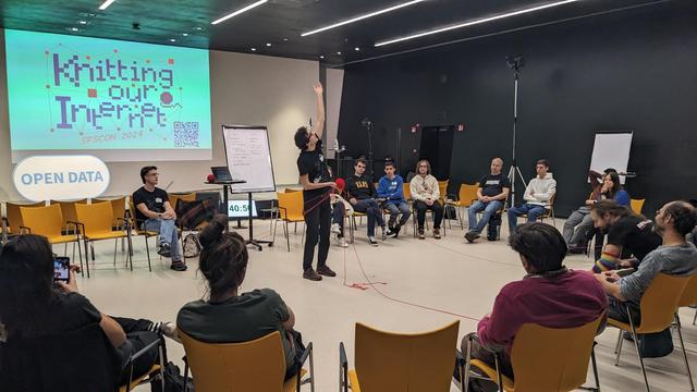 circle of chairs with people sitting there. I. the middle, person raising an arm. in the other hand he holds red threads that are connected with participants sitting on the chairs.