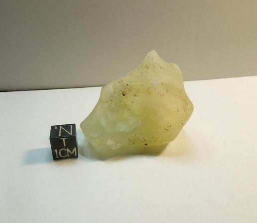 A small gemmy piece of Libyan Desert Glass next to a black centimeter scale cube.