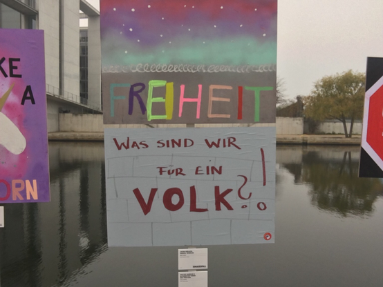 Zwei gemalte, beschriftete Schilder an der Spree, oben "Freiheit" in bunten Buchstaben vor Mauer mit Stacheldraht, unten die Frage "Was sind wir für ein Volk?“ in roter Schrift auf weißem Grund. Darunter kleine Tafeln mit den Urhebern: Oben Arndt-Gymnasium, unten Grips-Theater.