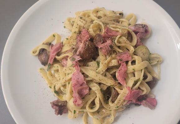 Farbfoto: Auf einem weißen Teller liegt eine Portion Bandnudeln mit gebratenen Champignons, ein paar halben Rosenkohlköpfen, Petersilie und einigen Streifen vom Galloway-Filet von gestern. Darüber ist ordentlich gemahlener Pfeffer. 