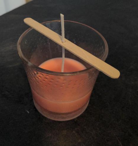 A salmon pink colored candle setting in a medium sized glass. A wooden posicle stick lays across the top of the glass to support the wick as the candle sets.