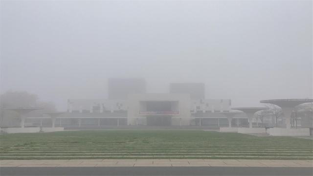 The Staatstheater Darmstadt, shrouded in fog, seen from Georg-Büchner-Platz on 11 November 2021. In the foreground, a terraced square with grass, on each side are several white, mushroom-like concrete objects leading to the barely visible Staatstheater Darmstadt in the background, a wide concrete building with a white, futuristic façade and two rectangular towers on top, which blend into a white sky. A banner above the entrance shows the theater’s season motto „Worauf hoffen?“ (What to hope for?)

Photo: Stefan Romero Grieser CC BY-SA 4.0