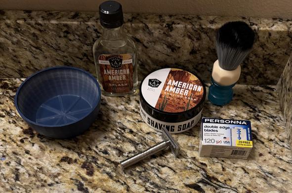 Cosmetic products on a countertop. Products include a shaving bowl, a shaving brush, a container of shaving soap, a bottle of aftershave, a double edge safety razor, and a package of double edge razor blades.