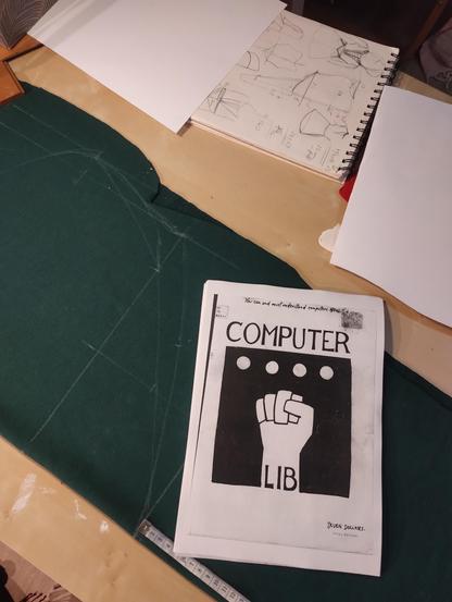 A table, on it a cape pattern drawn on green fabric and a book, titled Computer Lib, on top.