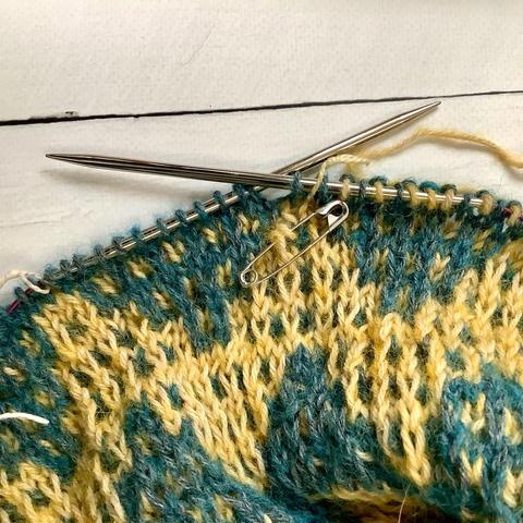 Closeup of a mosaic shawl in yellow and teal yarn. There are 2 loose stitches, caught with a safety pin.