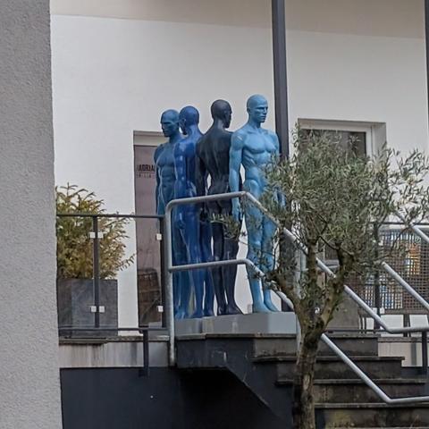 Vier nackte Schaufensterfiguren gefärbt in unterschiedlichen Blautönen stehen in verschiedene Richtungen gewandt auf einer Terrasse.