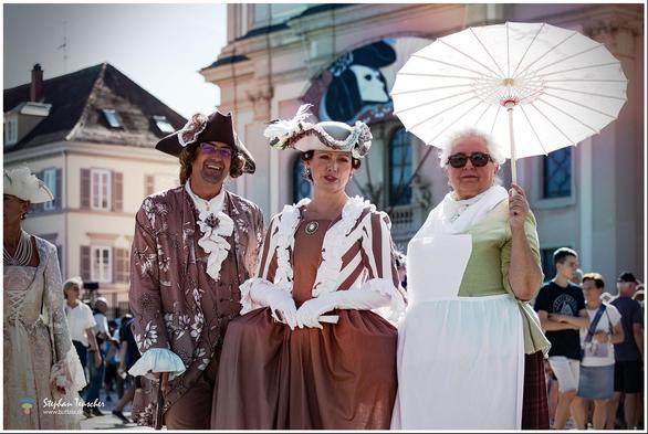 Das Foto zeigt drei Personen in historischen Kostümen. Ein Mann in brauner Kleidung mit Dreispitz, eine Frau im Kleid mit Rüschen und Hut, und eine Frau mit Schürze und Sonnenschirm. Der Hintergrund zeigt eine Kulisse mit klassischen Gebäuden.