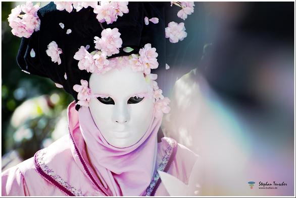 Das Bild zeigt eine maskierte Person mit einer weißen, ausdruckslosen Maske. Der schwarze Kopfschmuck ist mit rosa Blüten verziert. Ein rosa Schal und ein aufwendiges, besticktes Kostüm runden das mystische, sanfte Erscheinungsbild ab.