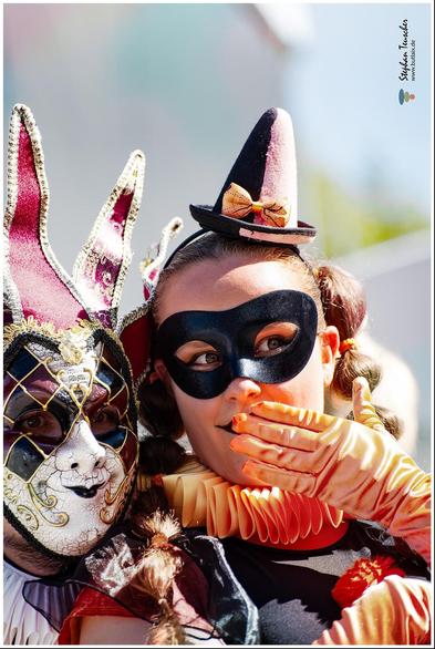 Das Foto zeigt zwei kostümierte Personen mit venezianischen Masken, bunten Kleidern und auffälligen Accessoires. Eine Person trägt ein Harlekin-Muster, die andere einen kleinen Hut und orange Handschuhe.