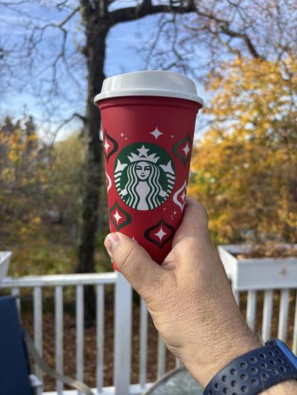 A Starbucks red holiday cup is held up against the background of fall foliage. 