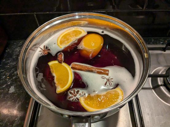 A large pan sitting on a stove. Red wine with orange quarters, cinnamon sticks, star anise and cloves floating in it. 