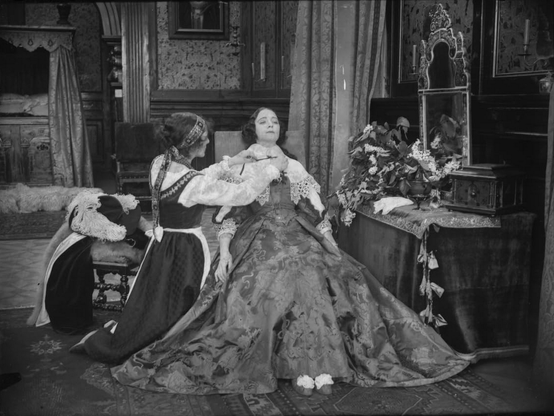 a woman seated in a chair in an elegantly furnished room, an attendant is  down on one knee, helping her to put on her fine clothing