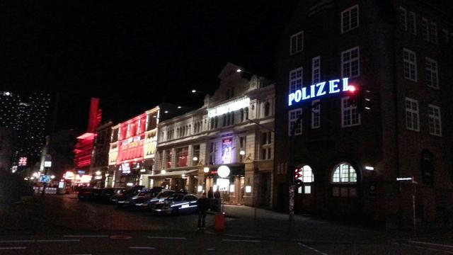 Bildbeschreibung von KI:
Das Foto zeigt die Davidwache auf der Reeperbahn in Hamburg bei Nacht. Die Davidwache ist eine der bekanntesten Polizeiwachen Deutschlands und ein Wahrzeichen im Stadtteil St. Pauli. 

Rechts im Bild befindet sich das Gebäude der Wache, das mit einem auffälligen, blauen "POLIZEI"-Leuchtschild markiert ist. Das Gebäude ist im traditionellen Backsteinstil gehalten und weist zahlreiche Fenster auf, die eine symmetrische Anordnung haben.

Links von der Davidwache erstreckt sich die Reeperbahn mit verschiedenen beleuchteten Gebäuden, einige davon in kräftigen roten und weißen Farben, was auf Clubs, Bars und andere Unterhaltungsbetriebe hindeutet. Die Szenerie ist charakteristisch für das bunte Nachtleben von St. Pauli, das sich entlang der Reeperbahn abspielt. Im Vordergrund sind einige parkende Autos und ein paar Fußgänger zu sehen, die das Bild zusätzlich beleben.