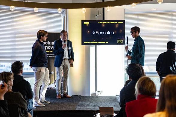Three men stand on stage, one holding a microphone as they interact with each other in front of an audience. The background screen displays "SensoTec," with a banner on the side that reads "Inclusive Innovations."
