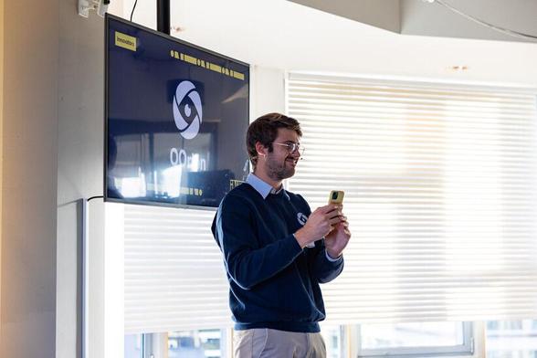 A man in a blue sweater holds a smartphone in both hands, smiling slightly while presenting. The screen in the background shows the logo "OOrion."
