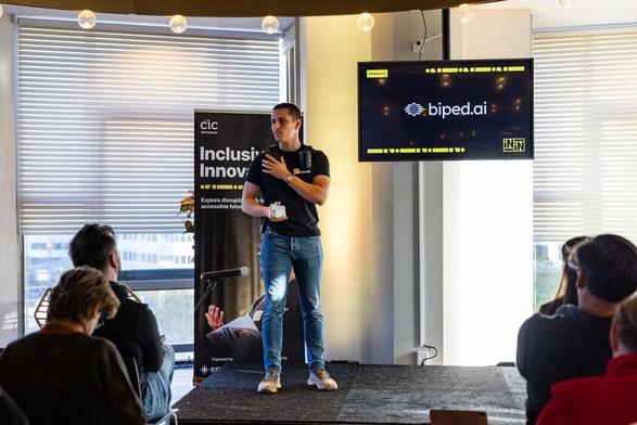 A man in a black T-shirt with a device strapped to his chest holds a small object while presenting. Behind him, the screen reads "biped.ai," with a banner that says "Inclusive Innovations."

