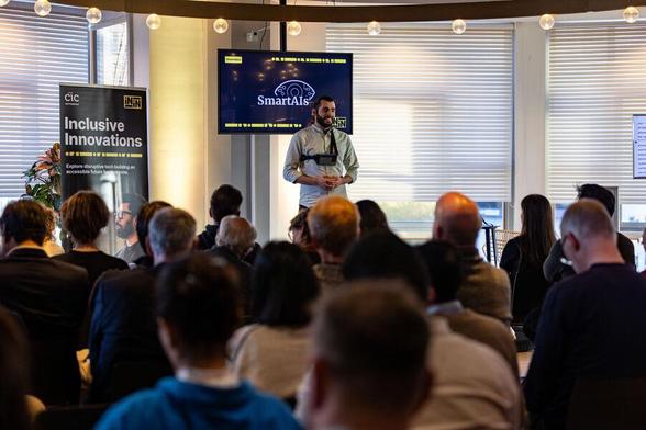 A man stands on stage, speaking in front of a screen that reads "SmartAIs." The audience is seated and facing him, with a banner to his left that reads "Inclusive Innovations."

