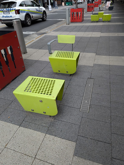 Photo of two bright green public chairs on the sidewalk. It's two small chairs instead of a big one. 