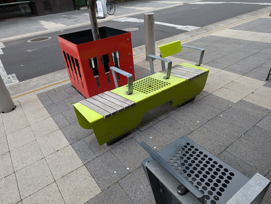 A bright green metal sitting place on the sidewalk. It's long enough for a person to sleep on, but it's got three metal armrests that look terribly uncomfortable as armrests because their true use is to not allow a person to sleep. 