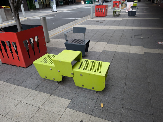 Photo of a bright green metal chair on the sidewalk. The body is long, but it has a huge chunk of metal in the middle. 