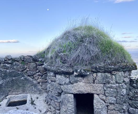 The moon tiny in a blue pre-dawn sky. In the foreground an ancient looking but well built stone structure, round and maybe 4 or 5 feet high including its peaked green turf roof. Well carved into bedrock next to it is a water trough. Beyond that you can see the stone wall that surrounds the area where the pigs would be kept. The whole thing looks like something out of a fantasy story, like where hobbits would keep pigs.