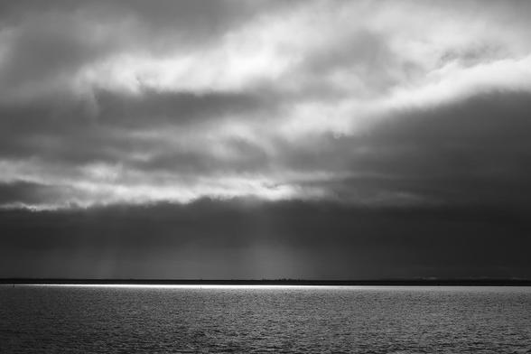 Photo of the sea with small waves. Above that a cloudy sky with sunlight coming through.
