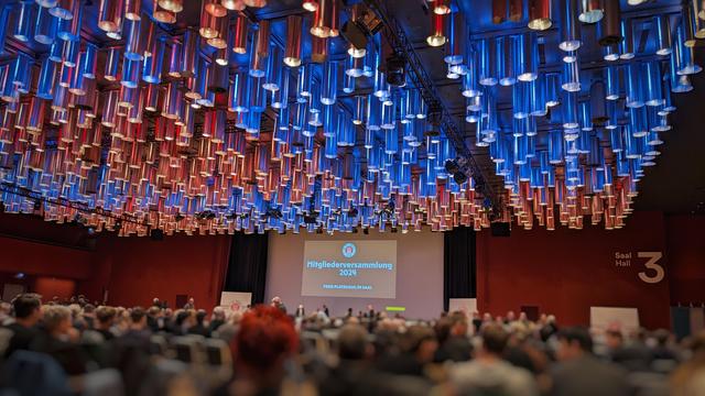 Bildbeschreibung von KI:
Das Foto zeigt den Innenraum von Saal 3 des CCH (Congress Center Hamburg) während der Mitgliederversammlung des FC St. Pauli im Jahr 2024. Im Vordergrund ist das Publikum zu sehen, das an der Veranstaltung teilnimmt, wobei die Köpfe der Teilnehmer unscharf sind. Im Hintergrund wird eine Leinwand mit der Aufschrift **"Mitgliederversammlung 2024"** und **"Freie Platzwahl im Saal"** projiziert.

Besonders auffällig ist die Decke des Saals, die mit einer Vielzahl von zylinderförmigen Lichtinstallationen gestaltet ist. Diese sind in Blau- und Rottönen beleuchtet, was eine dynamische und moderne Atmosphäre schafft. Rechts im Bild befindet sich die Beschriftung **"Saal Hall 3"** an der roten Wand, die den Veranstaltungsort kennzeichnet.