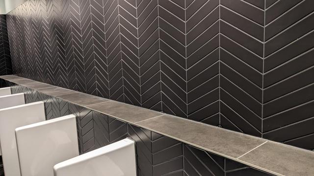Bildbeschreibung von KI:
Das Foto zeigt eine moderne, minimalistisch gestaltete Toilette im CCH Hamburg. Die Wand ist mit schwarzen Fliesen in einem Fischgrätenmuster gefliest, wobei die Fugen in Weiß kontrastieren und das Muster betonen. 

Unterhalb der gefliesten Wand verläuft eine Ablage aus grauem Stein oder Steinimitat, die flächenbündig und schlicht gestaltet ist. Darunter befinden sich mehrere weiße Urinale mit einem modernen, kantigen Design, die teilweise angeschnitten im unteren Bildbereich zu sehen sind. 

Die Atmosphäre ist klar und stilvoll, mit einem Fokus auf moderne Materialien und symmetrisches Design. Es herrscht ein sauberer und professioneller Eindruck.