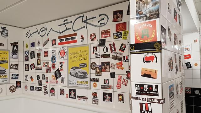 Bildbeschreibung von KI:
Das Foto zeigt eine Wand in einer Toilette des Millerntor-Stadions. Die Wand ist vollständig mit weißen Fliesen bedeckt, die dicht mit Aufklebern, Graffiti und anderen visuellen Elementen versehen sind. Der obere Bereich zeigt ein Graffiti mit der Aufschrift "HOWICK?", das in blauen Buchstaben geschrieben wurde.
Inhalte der Aufkleber:
Die Mehrheit der Aufkleber bezieht sich auf den FC St. Pauli, seine Fans und die ideologische Ausrichtung des Vereins:
"FORZA SANKT PAULI": Ein Slogan der Fanszene.
"Hamburg gegen Polizeigewalt": Ein sozialkritischer Aufkleber.
"ANTIFASCHISTISCH" mit Antifa-Logo: Zeigt die politische Haltung der Fans.
"You'll never rock alone": Eine humorvolle Abwandlung des bekannten Fußball-Slogans.
"Nik Gutfj" und "St. Pauli Lümmls": Weitere Beispiele für kreative Fangruppen-Aufkleber.
Aufkleber mit Totenkopfmotiv: Symbolisiert die Marke des Vereins.
- Diverse weitere Aufkleber mit Motiven wie Stadionbildern, sozialkritischen Aussagen, und Unterstützungsbekundungen für den FC St. Pauli.
Kommerzielle Elemente:
Ein gelber Werbeaufkleber für "Starcar Autovermietung" ist ebenfalls sichtbar, was einen Kontrast zu den überwiegend fanbezogenen und politischen Botschaften bildet.
Atmosphäre:
Die dichte Anordnung der Aufkleber und Graffiti vermittelt die typische Fan-Kultur und Individualität des Millerntor-Stadions. Die Botschaften repräsentieren sowohl die Leidenschaft der Fans als auch die politische und kulturelle Identität des Vereins.