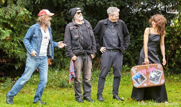 Ein Bandfoto vor einem Gebüsch. Drei Männer in Jeans, eine Frau mit einem Köfferchen im Abendkleid.