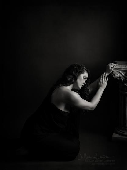 Toned black and white photo of model Jessa Ray Muse, torso wrapped in black fabric kneeling in front of a pillar.  A very low-key scene (greys on blacks to add an air of somberness.