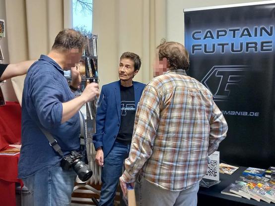 Das Bild zeigt rechts im Hintergrund den Futuremania-Stand auf der "EppiCon 1" und das schwarz-blaue Rollup mit dem Schriftzug „Captain Future“. Daneben, d. h. etwa in der Bildmitte, steht Website-Gründer und Betreiber Futuremania-Curtis alias Arne Sitzius (mit schwarzem Futuremania-T-Shirt und blauem Jackett), und rechts und links vor ihm Besucher der Veranstaltung, mit denen er gerade über Captain Future spricht...
