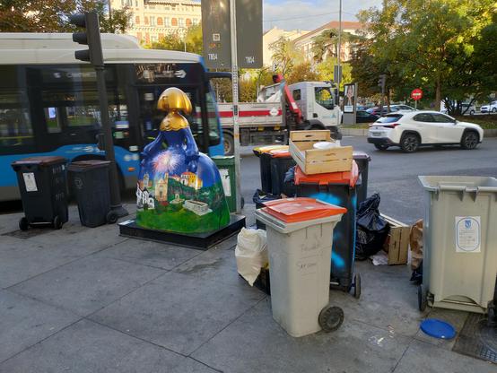 Una menina a tamaño natural en colores verde, azul y dorado. Está rodeada de cubos de basura y otros restos. De fondo el tráfico del paseo de Recoletos, un autobús, un camión y un coche blanco.