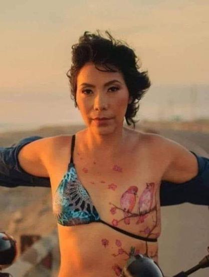 
Una mujer con el pelo corto y oscuro se encuentra al aire libre, con el sol poniéndose detrás de ella. Lleva un bikini azul y turquesa con un diseño intrincado. Sobre la cicatriz, de la mastectomía total de su pecho izquierdo a la vista, tiene un tatuaje  que representa dos pájaros posados ​​en una rama de cerezo en flor. Su expresión es seria. El fondo es borroso, mostrando un paisaje costero.