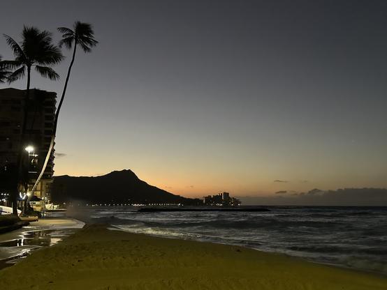A serene beach scene at dawn, featuring silhouettes of palm trees and a mountain in the background. The sky transitions from dark to a soft orange hue, while waves gently lap at the sandy shore. A faint city skyline is visible on the right, illuminated
