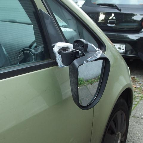 Rechter Außenspiegel eines geparkten Kleinwagens. Der Spiegel hängt im 90 Grad-Winkel mit der Spiegelfläche vom Fahrzeug wegzeigend herab und ist mit transparenter Plastikfolie notdürftig ›repariert‹.