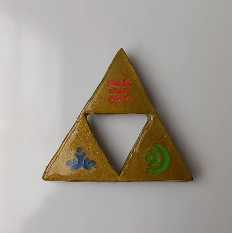 Imagen de un imán grande de la Trifuerza pegado a la pared: una figura dorada triangular compuesta por 3 triángulos dejando en el medio un triángulo invertido vacío. El triángulo superior tiene el símbolo de Din (3 líneas ondulantes rojas con un círculo en la parte derecha de la línea superior y otro en la parte izquierda de la línea inferior), el triángulo inferior izquierdo con el símbolo de Nayru (tres círculos azules formando un triángulo con una medialuna alrededor), y el triángulo inferior derecho con el símbolo de Farore (un círculo verde con dos medias lunas concéntricas).