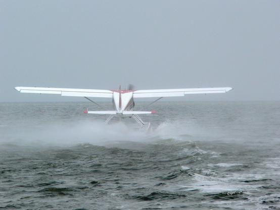 A De Havilland Canada DHC-2 Beaver seaplane is taking off from the water. The plane is seen from behind, with its floats (pontoons) skimming the surface of the water, creating a spray. The propeller is blurred, indicating motion. The background shows a vast expanse of water under a cloudy sky.