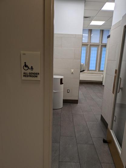 Photo of the entrance to a handicap-accessible gender neutral bathroom. There is a sign that states: "all gender bathroom." A trashcan, a wall-mounted hand drier and several opaque exterior windows are visible in a well-lit room.