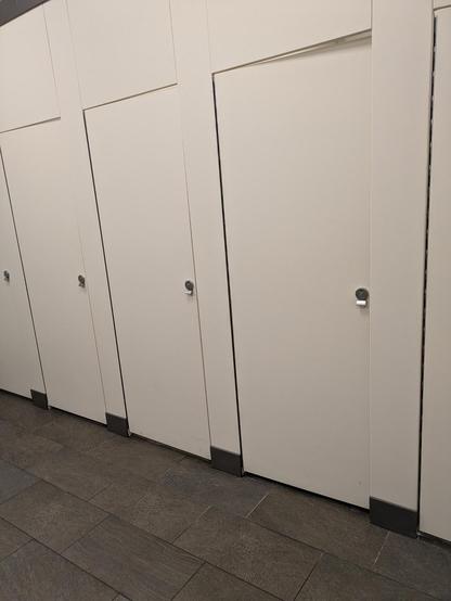 Photo of a row of white doors on a white wall. Each door marks a separate bathroom stall with an individual color-coded (red = occupied, green = empty) lock on the door.