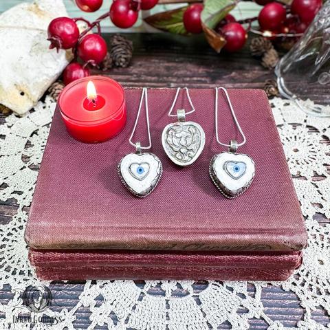 Three specimens of the "Heart Shaped Evil Eye Tibetan Necklace" product are arranged on a stack of old red-covered hard-bound books in front of a tealight candle. They are made up of white and blue stones in an Evil Eye Talisman shape mounted in a silver heart-shaped bezel. Presented by Inked Goddess Creations.