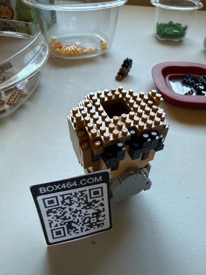 A small Lego figure almost complete. There is a small hole on top of the head. A printed qrcode sits next to it, which links to my profile. 