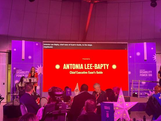A red screen on a stage lit by puple lighting reading Antonia Lee-Bapty Chief Executive Euan's Guide. There are people sitting in front of the screen and stage. 