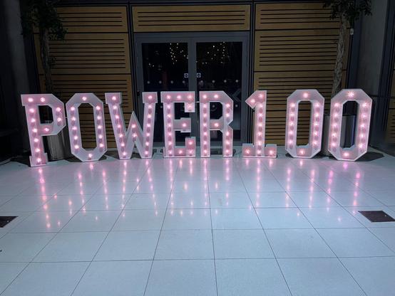 A photo of light up letters reading Power 100 on a white tiled floor. 