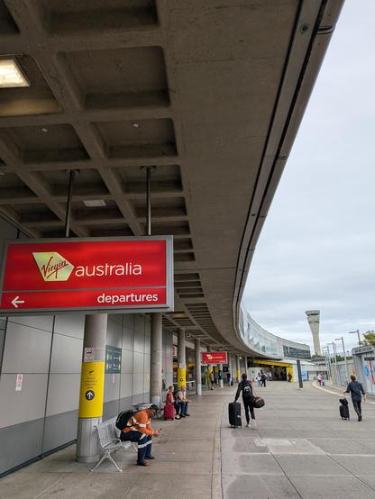The outside of the Brisbane Domestic Airport terminal with the Virgin Australia red departures sign in view. The gentle curve of the building arcs away with the air control tower at its end, clearly visible against a cloudy sky.
