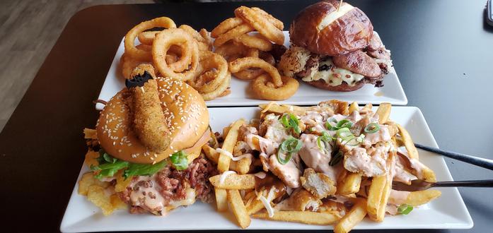 Two rectangular plates with food. The closer one has a burger on the left with a side of poutine to the right. The plate behind it has a burger on the right with a side of onion rings to it's left.