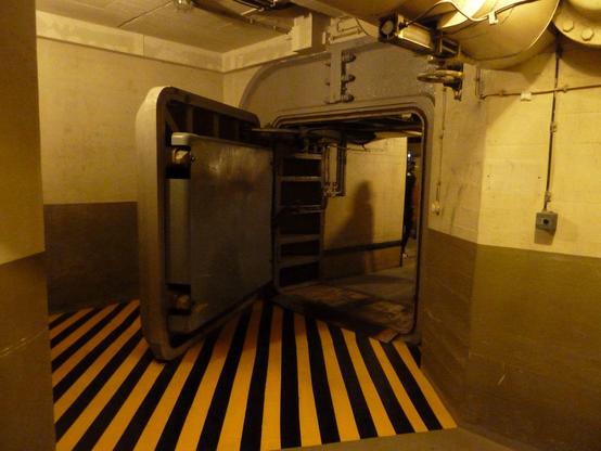 Eine schwere, dicke Strahlenschutztür, die geöffnet ist. Auf dem Boden schwarz-gelbe Streifen, die den Bereich markieren, in dem man sich im Alarmfall nicht aufhalten darf. Die Wände sind Sichtbeton, das untere Drittel grau, der obere Teil schmutzigweiß. Auf den Wänden sind verschiedene Leitungen, z. B. Kabel zu den Lichtschaltern etc. zu sehen. Das Bild ist insgesamt etwas dunkel (und auch leicht verwackelt), was den schlechten Lichtverhältnissen geschuldet ist.