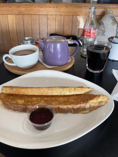 Two slices of French Toasr on a white plate. With a small tub of maple syrup. With a glass of Coca Cola and a cup of tea beside the plate. 